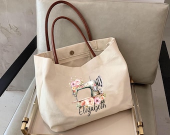May include: Beige canvas tote bag with brown leather handles. The bag features a floral design with a vintage sewing machine and the name "Elizabeth" printed in black. The tote is sitting on a gold-trimmed tray.