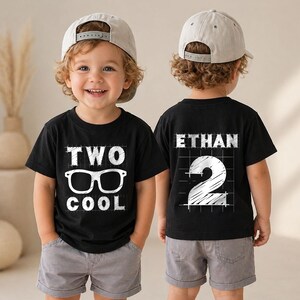 May include: A black t-shirt featuring white graphic text that reads TWO COOL with an illustration of glasses on the front, and the name ETHAN with the number 2 on the back, paired with grey shorts and a light grey baseball cap worn by a toddler.