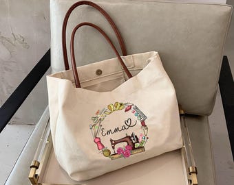 May include: A beige canvas tote bag with brown leather handles. The bag features a circular design with sewing-related illustrations and the name "Emma". The design includes a sewing machine, scissors, and a heart.
