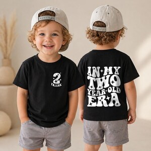 May include: A black t-shirt featuring a white graphic on the front left chest with the number 2 and the name Frank. The back of the shirt displays the white text In My Two Year Old Era with star accents. The shirt is paired with grey shorts.