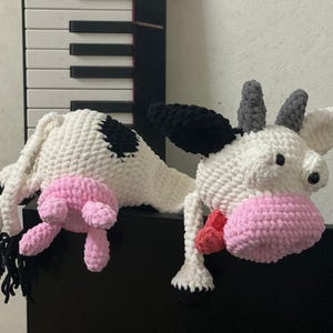 Op de afbeelding: Een gehaakt knuffeldier in de vorm van een koe met zwarte en witte vlekken. De koe heeft roze oren en een roze neus. Het speelgoed ligt op een zwart oppervlak.