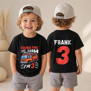 May include: Two black t-shirts with a fire truck graphic and the text "SOUND THE ALARM I'M 3" on the front, and the name "FRANK" above a large red number "3" on the back. Both shirts are paired with grey shorts and light-colored baseball caps.