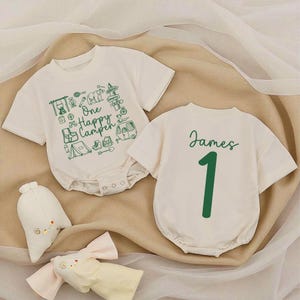 May include: Two off-white baby onesies. One has a green graphic with camping-themed icons and the text "One Happy Camper." The other has the name "James" and the number "1" in green. Two stuffed toys are in the foreground.