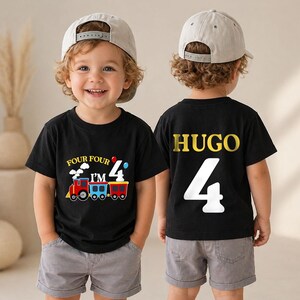 May include: A black t-shirt featuring a colorful train graphic with the text 'FOUR FOUR I'M 4' on the front and the name 'HUGO' above a large white number '4' on the back, paired with gray shorts and a light gray baseball cap worn by a child.