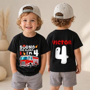 May include: A black t-shirt featuring a red fire truck graphic, a fire hydrant, flames, and the text 'SOUND THE ALARM I'M 4' on the front. The back of the shirt displays the name 'VICTOR' in red letters above a large white number 4.