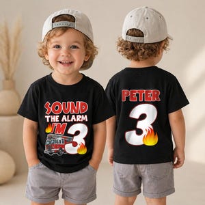 May include: A black t-shirt featuring the text SOUND THE ALARM I'M 3 with a fire truck graphic and flames, and a second black t-shirt displaying the name PETER above a large number 3 with flames. Both shirts are paired with light gray shorts and caps.