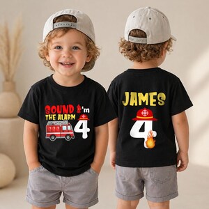 May include: A black t-shirt featuring a red fire truck graphic, the text 'SOUND THE ALARM', a fire hydrant, and the number 4. The back displays the name 'JAMES' in yellow, a red firefighter helmet, the number 4, and a small orange flame graphic.
