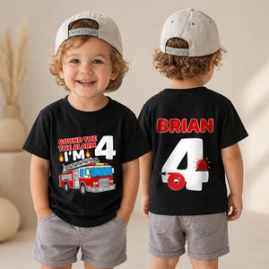 May include: Two black t-shirts featuring a fire truck theme. The front displays a red fire truck with the text 'SOUND THE ALARM I'M 4'. The back features the name 'BRIAN' above a large white number 4 with a red fire hose and alarm light.