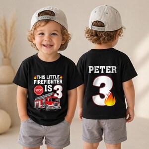 May include: A black t-shirt featuring a red fire truck graphic, a stop sign, and the text This Little Firefighter Is 3 on the front, with the name Peter and the number 3 with a flame graphic printed on the back, worn with gray shorts.