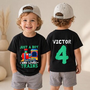 May include: A black t-shirt featuring a colorful train graphic with the text "JUST A BOY WHO LOVES TRAINS" on the front and the name "VICTOR" above a large green number "4" on the back, paired with grey shorts and a light beige baseball cap.