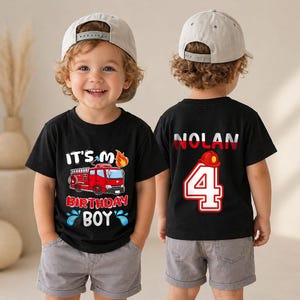 May include: A black t-shirt featuring a red fire truck graphic with the text IT'S MY BIRTHDAY BOY on the front and the name NOLAN with the number 4 and a small red fire helmet graphic on the back, paired with light gray shorts and a beige cap.