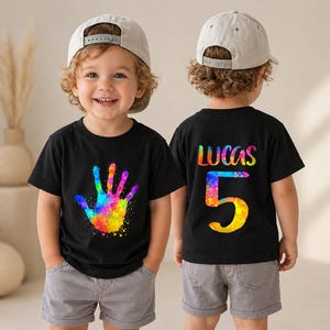 May include: A black t-shirt featuring a vibrant, multi-colored paint splatter handprint on the front. The back of the shirt displays the name LUCAS in colorful letters above a large, paint-splattered number 5. The shirt has short sleeves.