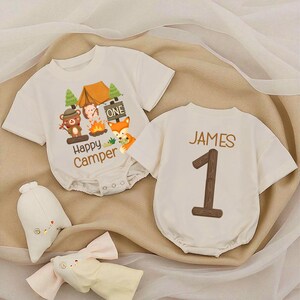 May include: Two off-white baby rompers. One features a camping scene with a bear, hedgehog, and fox, the text "Happy Camper" and "ONE". The other has the name "JAMES" and the number "1" printed on the back. Two stuffed animal toys are in the foreground.