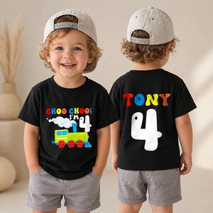 May include: A black t-shirt featuring a colorful train graphic with the text CHOO CHOO! I'M 4 on the front and the name TONY above a large white number 4 on the back, paired with gray shorts and a light gray baseball cap worn by a young child.