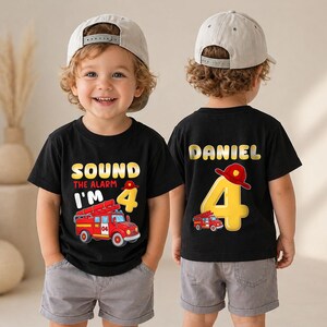 May include: A black t-shirt featuring a red fire truck graphic, the text "SOUND THE ALARM I'M 4" and a yellow number 4. The back of the shirt displays the name "DANIEL" above a large yellow number 4 with a red firefighter helmet.