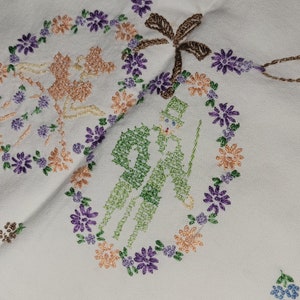 May include: White linen tablecloth with embroidered floral designs and a couple in a wreath. The embroidery is in shades of purple, pink, green, and brown.