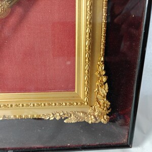 May include: A gold-colored picture frame with ornate detailing and a red velvet mat. The frame is made of wood and has a glass front.