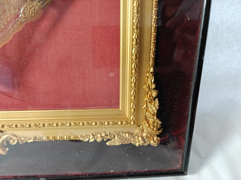 May include: A gold-colored picture frame with ornate detailing and a red velvet mat. The frame is made of wood and has a glass front.
