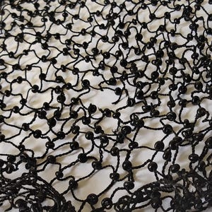 May include: Close-up of a black beaded net. The net is made of black thread and small, round black beads. The beads are evenly spaced throughout the net, creating a delicate and intricate pattern. The background is a light, off-white color.