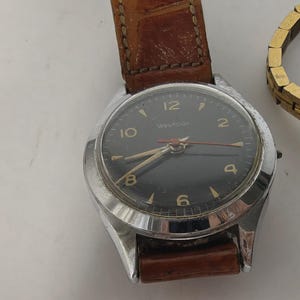 May include: A Westclox wristwatch with a round silver-tone metal case and a black dial featuring gold-tone numerals and hands. The watch is attached to a brown textured leather strap with visible stitching. A gold-tone metal link bracelet is nearby.