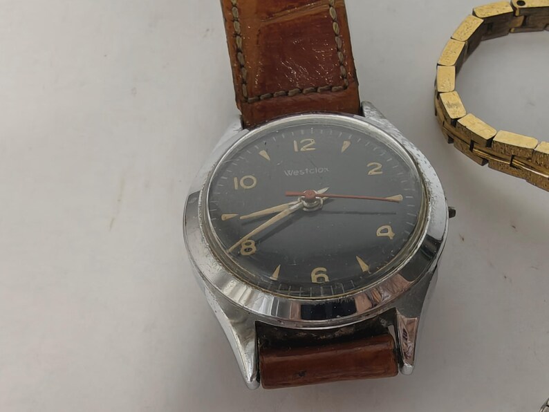 May include: A Westclox wristwatch with a round silver-tone metal case and a black dial featuring gold-tone numerals and hands. The watch is attached to a brown textured leather strap with visible stitching. A gold-tone metal link bracelet is nearby.