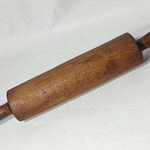 May include: A cylindrical wooden rolling pin with a dark brown finish and visible wood grain texture. The tool features a central barrel section and two smaller, rounded handles on each end, showing signs of wear and natural wood markings.