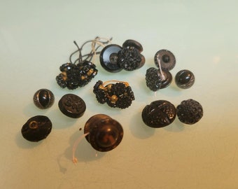 May include: A collection of various black buttons with different designs and textures. Some buttons have floral patterns, while others are smooth or have intricate detailing. Several buttons are attached to thread.