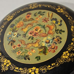 May include: A round, black decorative box with a floral and squirrel design. The center features a light green background with colorful flowers and squirrels. The rim is adorned with gold floral accents.