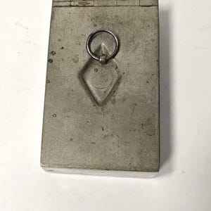 May include: A rectangular, weathered silver-toned metal box featuring a hinged lid and a central diamond-shaped indentation. A small, circular metal ring pull is attached to the center of the diamond-shaped detail on the top surface.