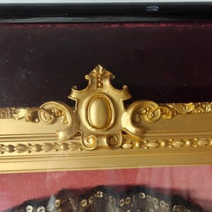 May include: A gold-colored ornate frame with a burgundy velvet mat. The frame has a decorative design with a central oval shape.