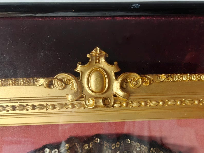 May include: A gold-colored ornate frame with a burgundy velvet mat. The frame has a decorative design with a central oval shape.