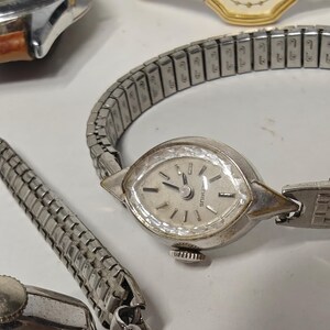 May include: A silver-tone wristwatch with a marquise-shaped case and a faceted crystal bezel. The watch features a light-colored dial with silver markers and a flexible, expandable metal link bracelet. The brand name Henrus is printed on the dial.