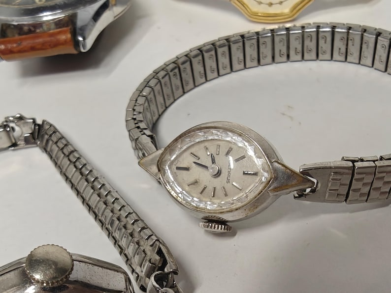 May include: A silver-tone wristwatch with a marquise-shaped case and a faceted crystal bezel. The watch features a light-colored dial with silver markers and a flexible, expandable metal link bracelet. The brand name Henrus is printed on the dial.