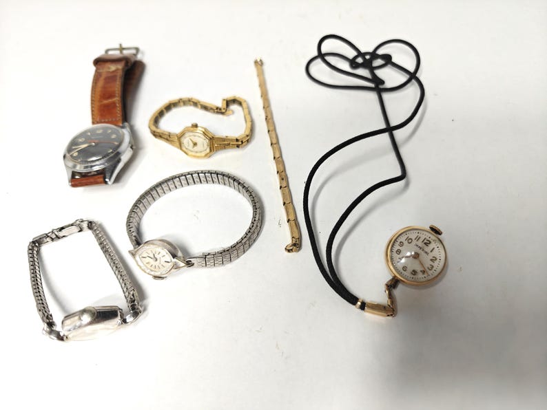 May include: A collection of timepieces including a watch with a brown leather strap, a gold-tone watch with a link bracelet, a silver-tone watch with an expansion band, a separate silver-tone watch band, a gold-tone chain, and a pendant watch on a cord.