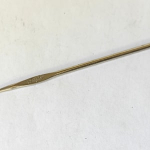 May include: A silver-colored crochet hook. The hook has a slender, elongated shaft with a curved hook at one end and a slightly wider handle with engraved text. The tool is designed for creating fabric from yarn.