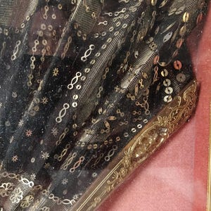 May include: A black fabric fan with gold sequins and a gold metal handle. The fan is framed and displayed on a pink background.