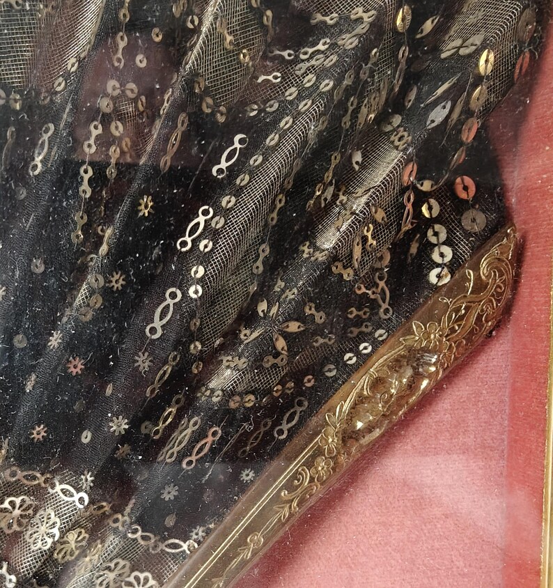 May include: A black fabric fan with gold sequins and a gold metal handle. The fan is framed and displayed on a pink background.