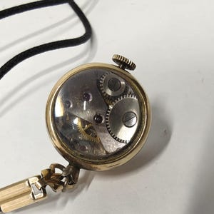 May include: A gold-toned round pendant watch with a clear glass back revealing the internal mechanical movement, including silver gears and small red jewels. The watch is attached to a thin black cord with a gold-toned metal connector piece.