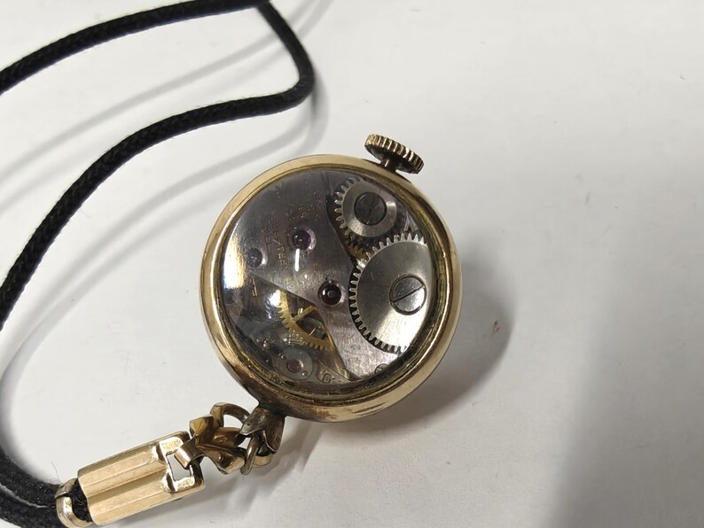 May include: A gold-toned round pendant watch with a clear glass back revealing the internal mechanical movement, including silver gears and small red jewels. The watch is attached to a thin black cord with a gold-toned metal connector piece.