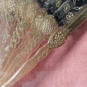 May include: A close-up of a vintage hand fan with a translucent, ornate design. The fan is made of a clear material with gold accents and intricate patterns. The fan is closed and the handle is visible.