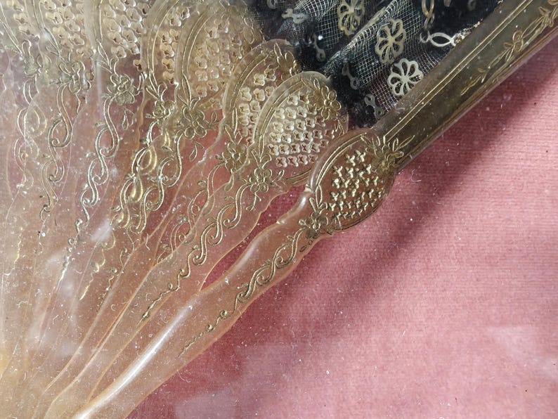 May include: A close-up of a vintage hand fan with a translucent, ornate design. The fan is made of a clear material with gold accents and intricate patterns. The fan is closed and the handle is visible.