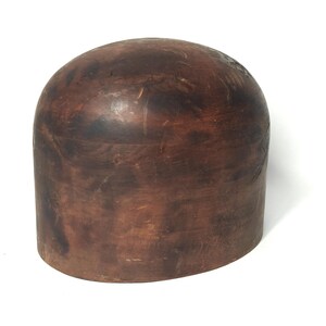 May include: A vintage, dome-shaped hat block made of dark brown wood. The hat block has a cylindrical base and a rounded top, showing the natural wood grain and variations in color. The surface has a smooth, aged appearance.
