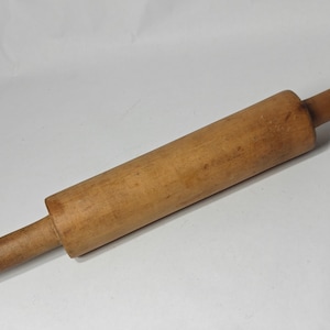 May include: A cylindrical wooden rolling pin with a natural light brown finish and visible wood grain. The kitchen tool features a central barrel section and two smaller, rounded handles on each end, showing signs of wear and use.