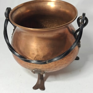 May include: A round copper cauldron with a hammered texture, featuring a wide open top and a dark, twisted metal swing handle. The vessel is supported by three dark metal feet attached to the base, providing a stable foundation for the pot.