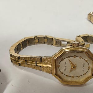 May include: An Armitron quartz wristwatch with an octagonal gold-tone case and a textured gold-tone link bracelet. The watch face is off-white with gold-tone dot hour markers and gold-tone hands. The word Armitron is printed on the dial.