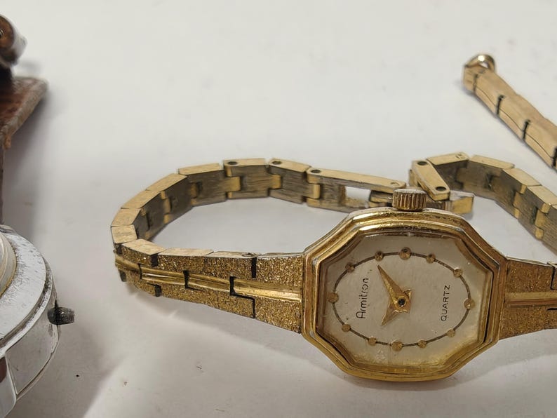May include: An Armitron quartz wristwatch with an octagonal gold-tone case and a textured gold-tone link bracelet. The watch face is off-white with gold-tone dot hour markers and gold-tone hands. The word Armitron is printed on the dial.