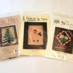 May include: Three embroidery kits with white backgrounds and colorful designs. The first kit is called "Pine Tree" and features a green pine tree on a red background. The second kit is called "A Stitch In Time" and features a black and white chicken on a pink background. The third kit is called "The Sweetheart Tree" and features two white flowers on a black background.