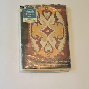 May include: A plastic-wrapped package containing a rug hooking pattern titled "Colonial Rug Pattern" by Amish Hearts. The pattern features a brown, yellow, and cream rug design with a heart motif. The package includes instructions for using Aunt Lydia's Heavy Rug Yarn and Punch Needle. The finished size of the rug is 24x30 inches.