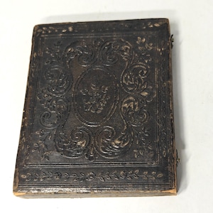 May include: A small, antique, dark brown leather-bound book with ornate embossed floral and scrollwork designs. The cover features a central oval with a floral motif and a decorative border. The edges show signs of wear.