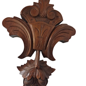 May include: A carved wooden decorative wall sconce with a dark brown finish. The sconce features a central vase-like element, flanked by curved, leaf-like designs. The bottom has a cluster of acorns and leaves.